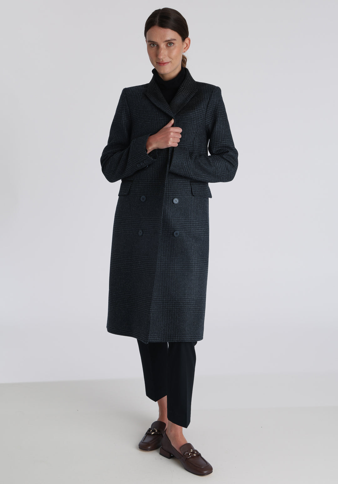 Model wearing the Cully coat in black and charcoal grey Prince of Wales check wool, a knee-length, double-breasted style with pointed lapels and a structured, straight-cut silhouette designed to flatter.