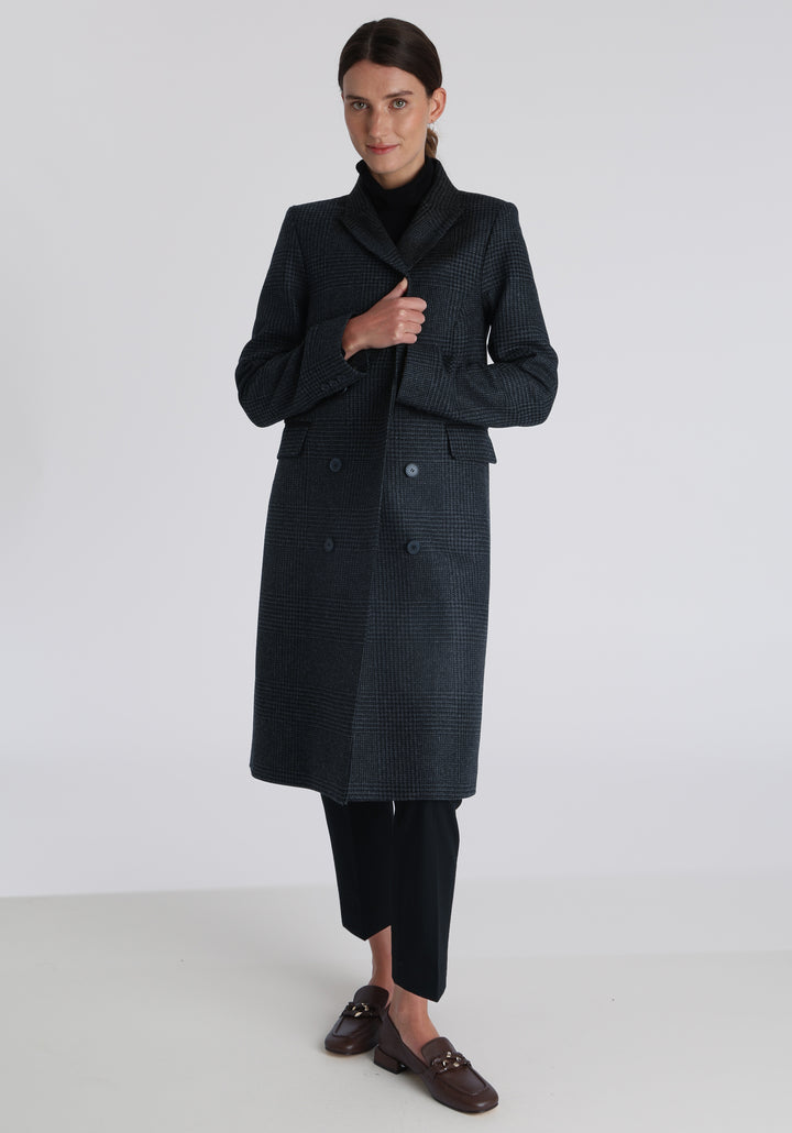 Model wearing the Cully coat in black and charcoal grey Prince of Wales check wool, a knee-length, double-breasted style with pointed lapels and a structured, straight-cut silhouette designed to flatter.