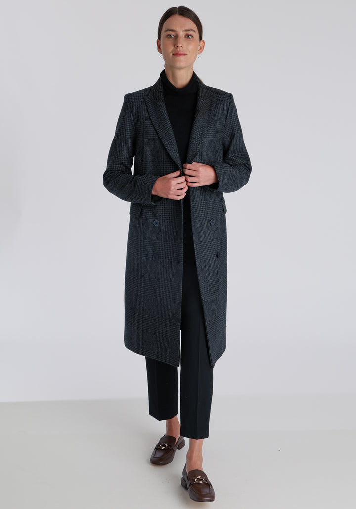 Model wearing the Cully coat in black and charcoal grey Prince of Wales check wool, a knee-length, double-breasted style with pointed lapels and a structured, straight-cut silhouette designed to flatter.