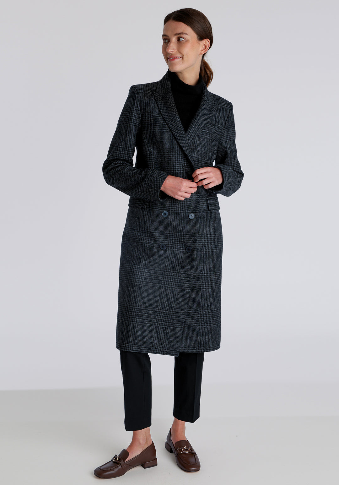 Model wearing the Cully coat in black and charcoal grey Prince of Wales check wool, a knee-length, double-breasted style with pointed lapels and a structured, straight-cut silhouette designed to flatter.
