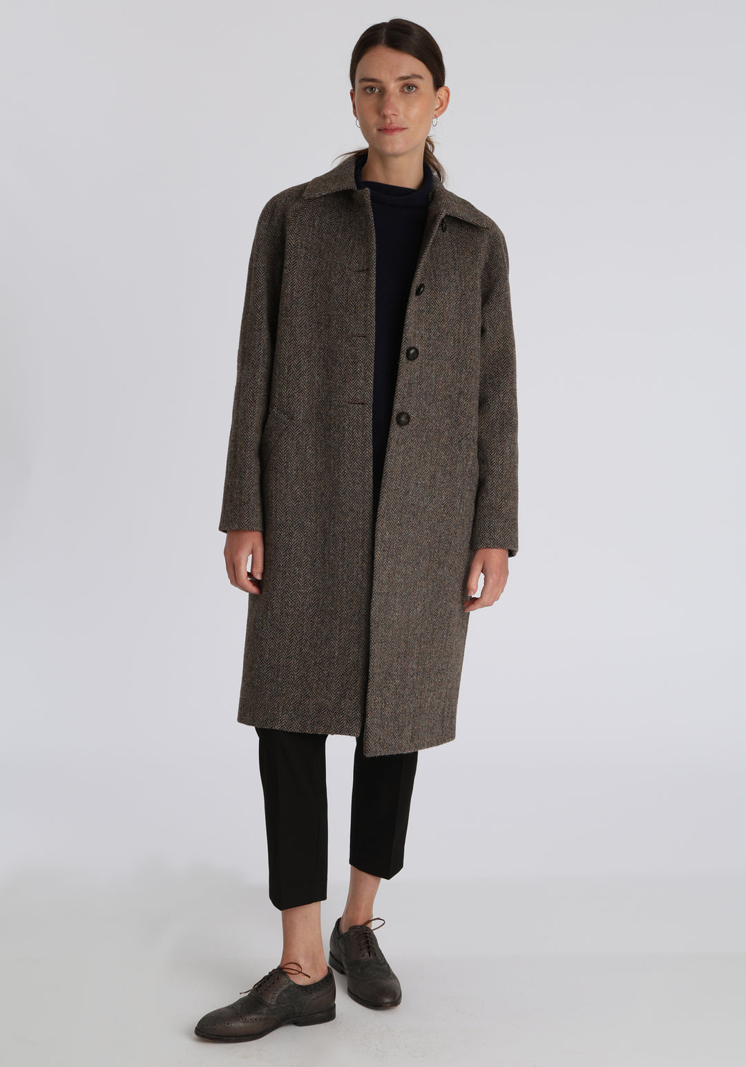 Model wearing the Dari coat in heather herringbone Harris Tweed, a relaxed cocoon-shaped style with raglan sleeves, finished with leather trim.
