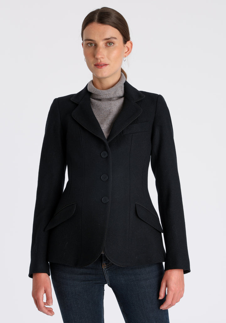 Model wearing the Hacking jacket in black textured merino wool, a slim fit style with notched lapels, angled hip pockets, and a single back vent, inspired by traditional equestrian tailoring.