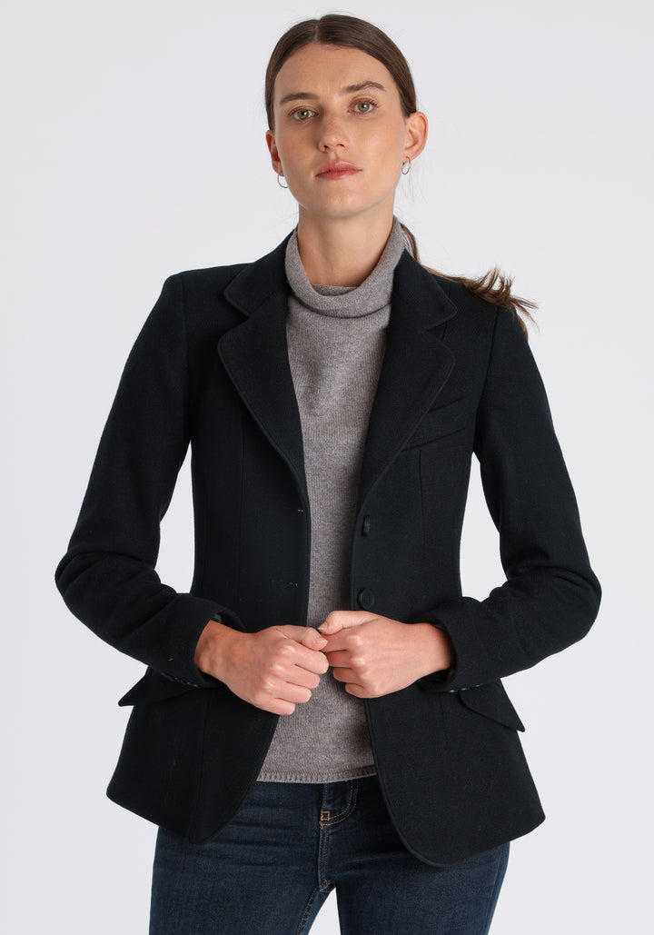 Model wearing the Hacking jacket in black textured merino wool, a slim fit style with notched lapels, angled hip pockets, and a single back vent, inspired by traditional equestrian tailoring.