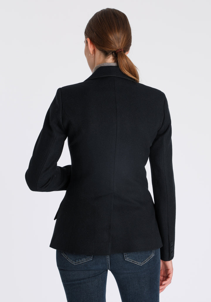 Model wearing the Hacking jacket in black textured merino wool, a slim fit style with notched lapels, angled hip pockets, and a single back vent, inspired by traditional equestrian tailoring.