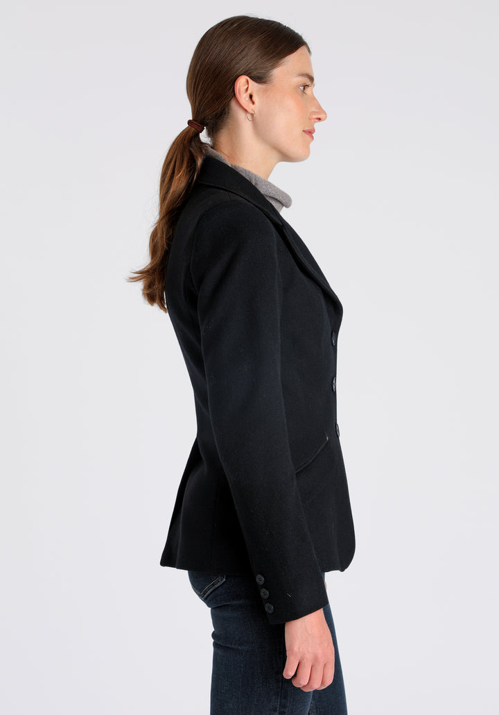 Model wearing the Hacking jacket in black textured merino wool, a slim fit style with notched lapels, angled hip pockets, and a single back vent, inspired by traditional equestrian tailoring.