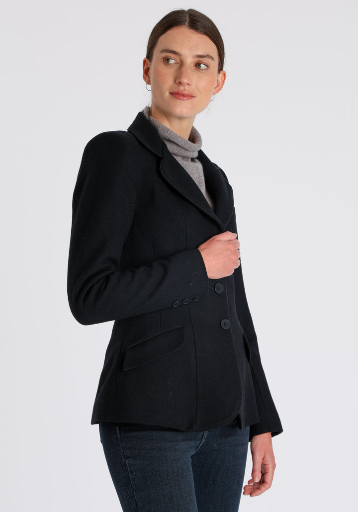 Model wearing the Hacking jacket in black textured merino wool, a slim fit style with notched lapels, angled hip pockets, and a single back vent, inspired by traditional equestrian tailoring.