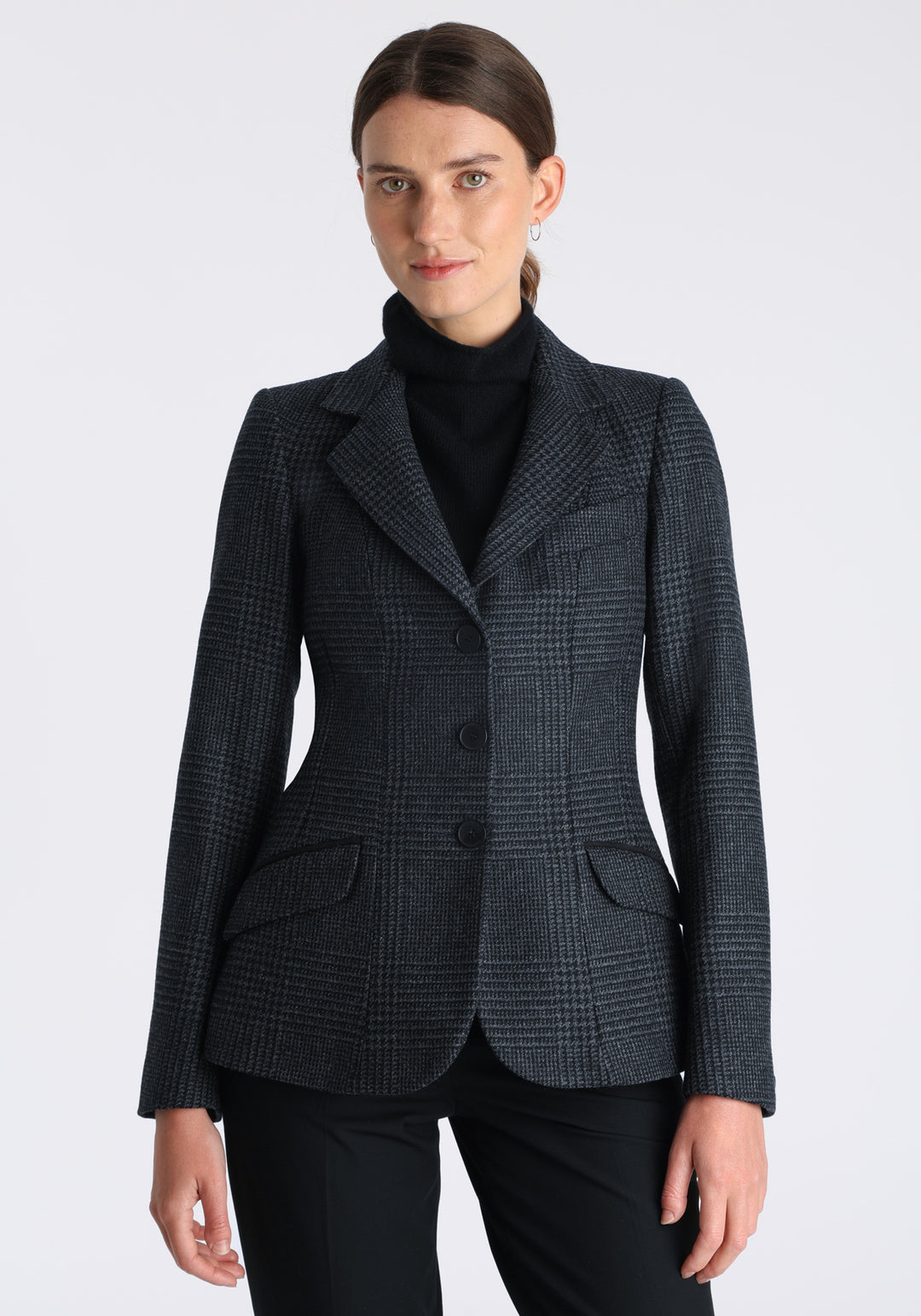 Model wearing the Hacking jacket in black and grey Prince of Wales check wool, a slim fit style with notched lapels, angled hip pockets, and a single back vent, inspired by traditional equestrian tailoring.