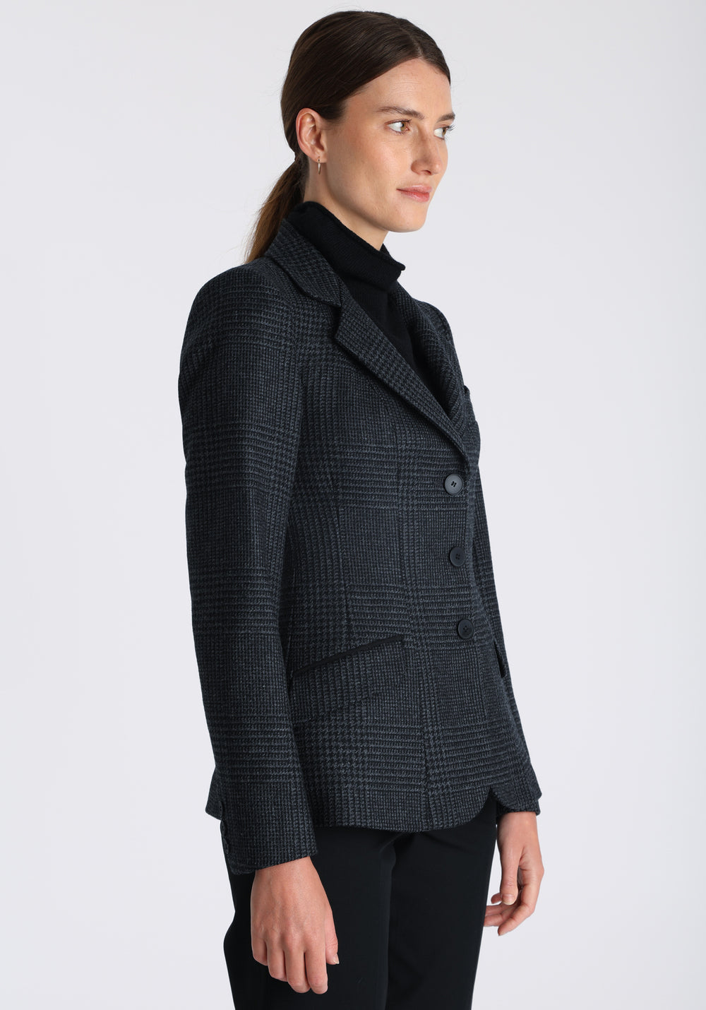 Model wearing the Hacking jacket in black and grey Prince of Wales check wool, a slim fit style with notched lapels, angled hip pockets, and a single back vent, inspired by traditional equestrian tailoring.