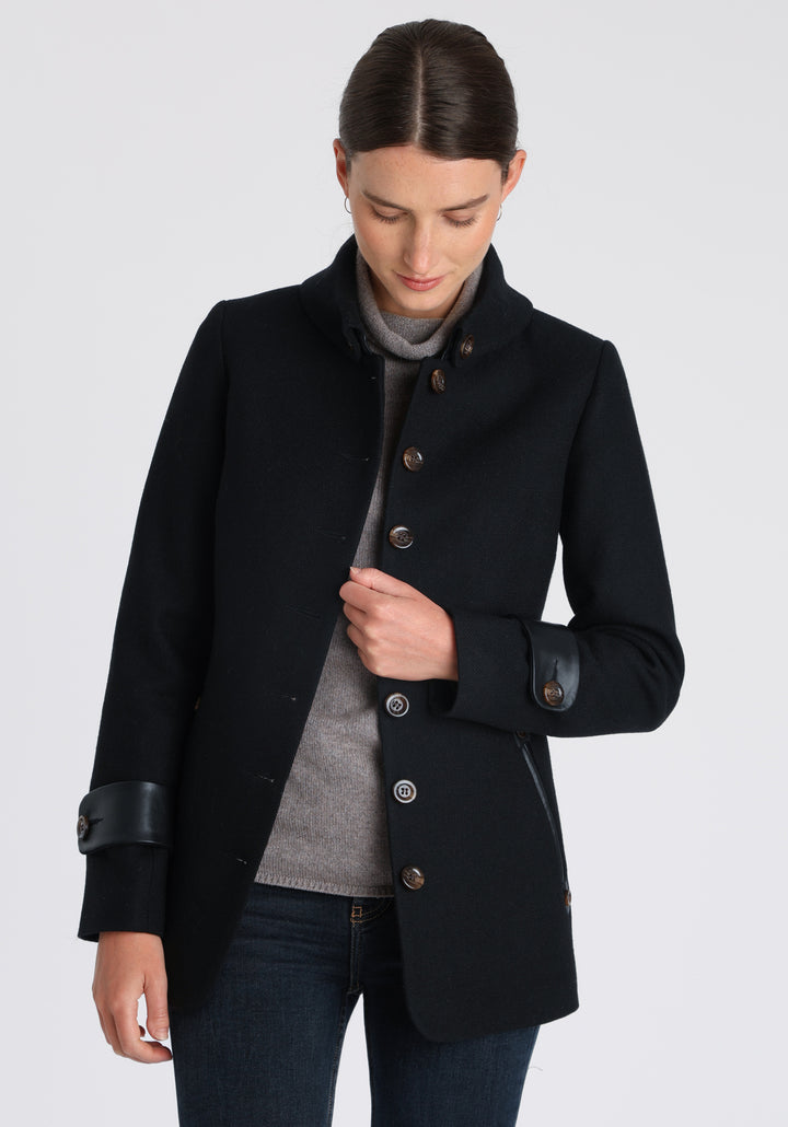 Model wearing the Hendre jacket in black textured wool, a structured style with a high collar, reversible leather cuff bands, and a back belt for versatile styling.