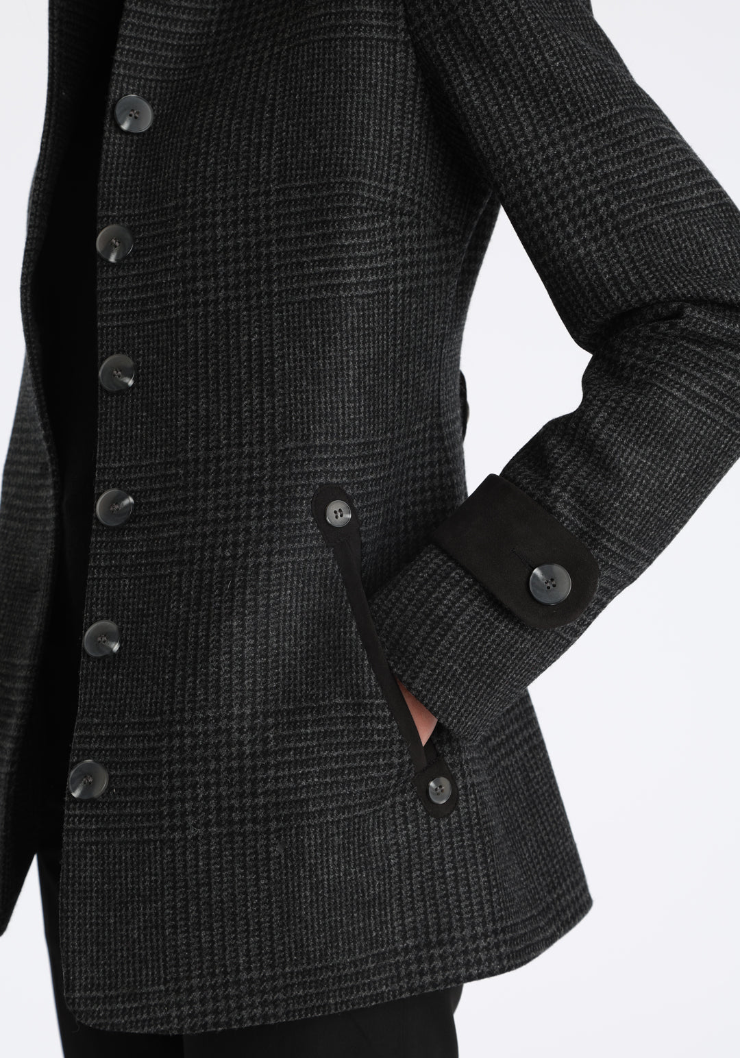 Model wearing the Hendre jacket in black and grey Prince of Wales check, a structured style with a high collar, reversible suede cuff bands, and a back belt for versatile styling.