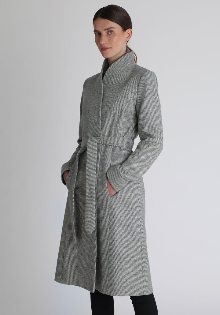 Model wearing the Hillsy coat in slate blue and cream lambswool twill, a wrap-style design with a standing collar and tie belt, offering a softly tailored, elegant silhouette. 