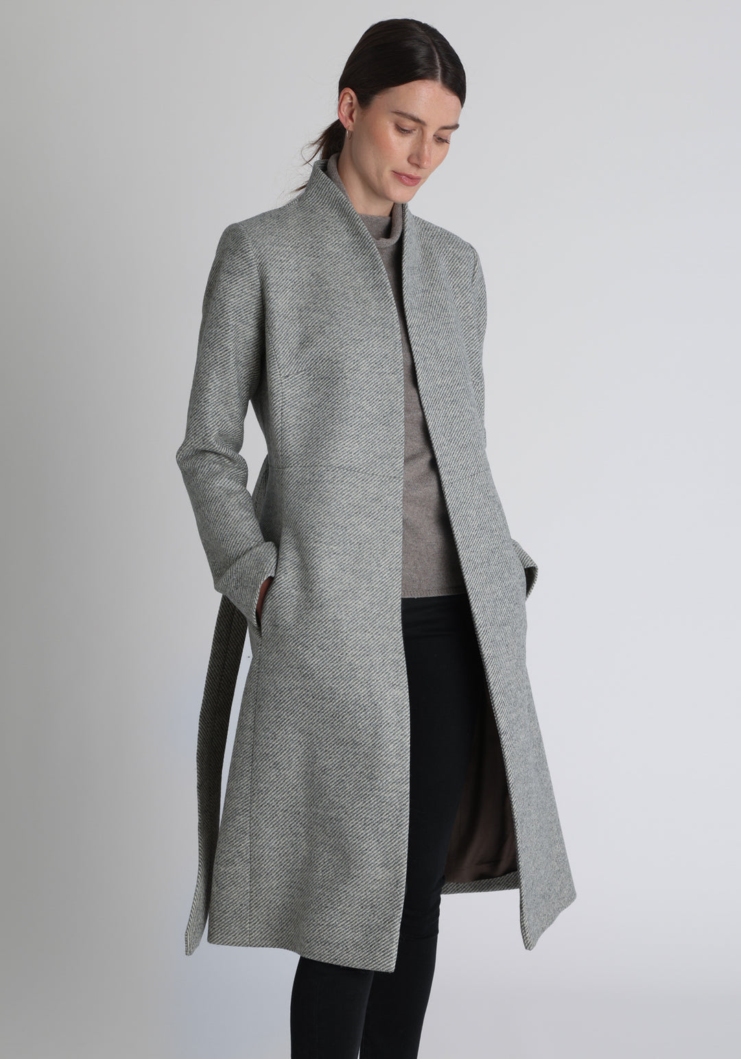 Model wearing the Hillsy coat in slate blue and cream lambswool twill, a wrap-style design with a standing collar and tie belt, offering a softly tailored, elegant silhouette. 