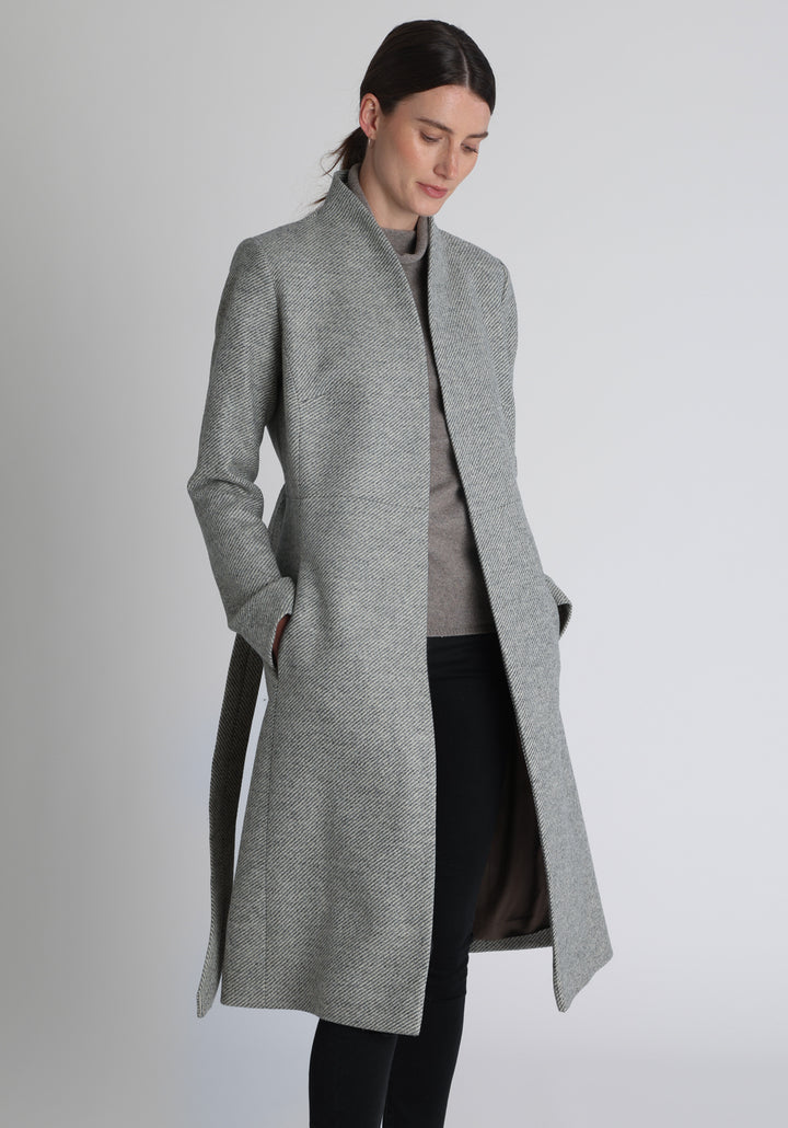 Model wearing the Hillsy coat in slate blue and cream lambswool twill, a wrap-style design with a standing collar and tie belt, offering a softly tailored, elegant silhouette. 