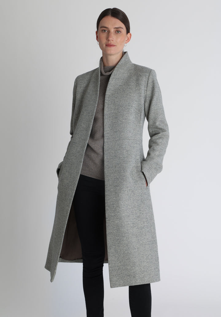 Model wearing the Hillsy coat in slate blue and cream lambswool twill, a wrap-style design with a standing collar and tie belt, offering a softly tailored, elegant silhouette. 
