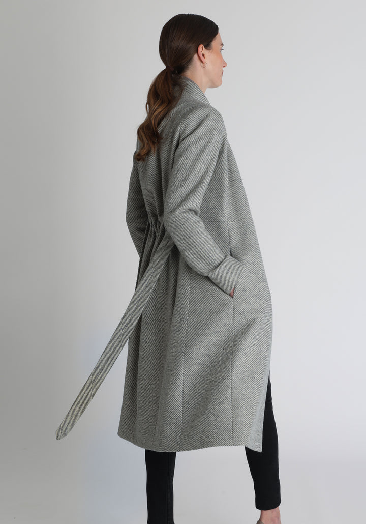 Model wearing the Hillsy coat in slate blue and cream lambswool twill, a wrap-style design with a standing collar and tie belt, offering a softly tailored, elegant silhouette. 