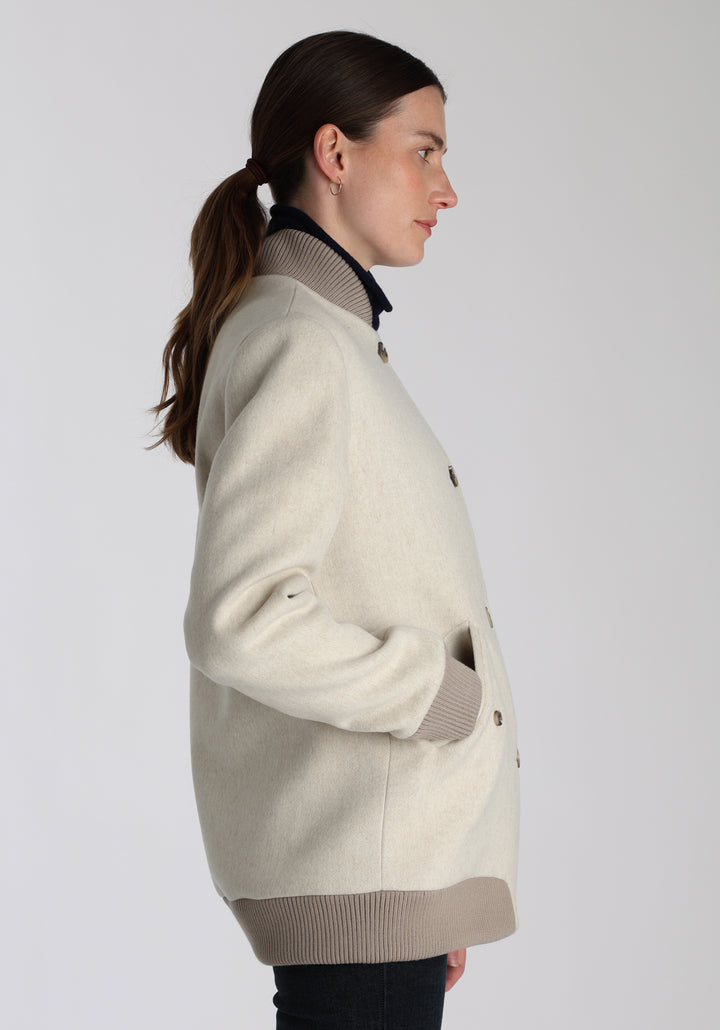 Model wearing the Jeffy jacket in cream merino wool, a baseball-inspired style with curved pocket flaps, front button fastenings, and ribbed knit trim, cut in a streamlined silhouette.