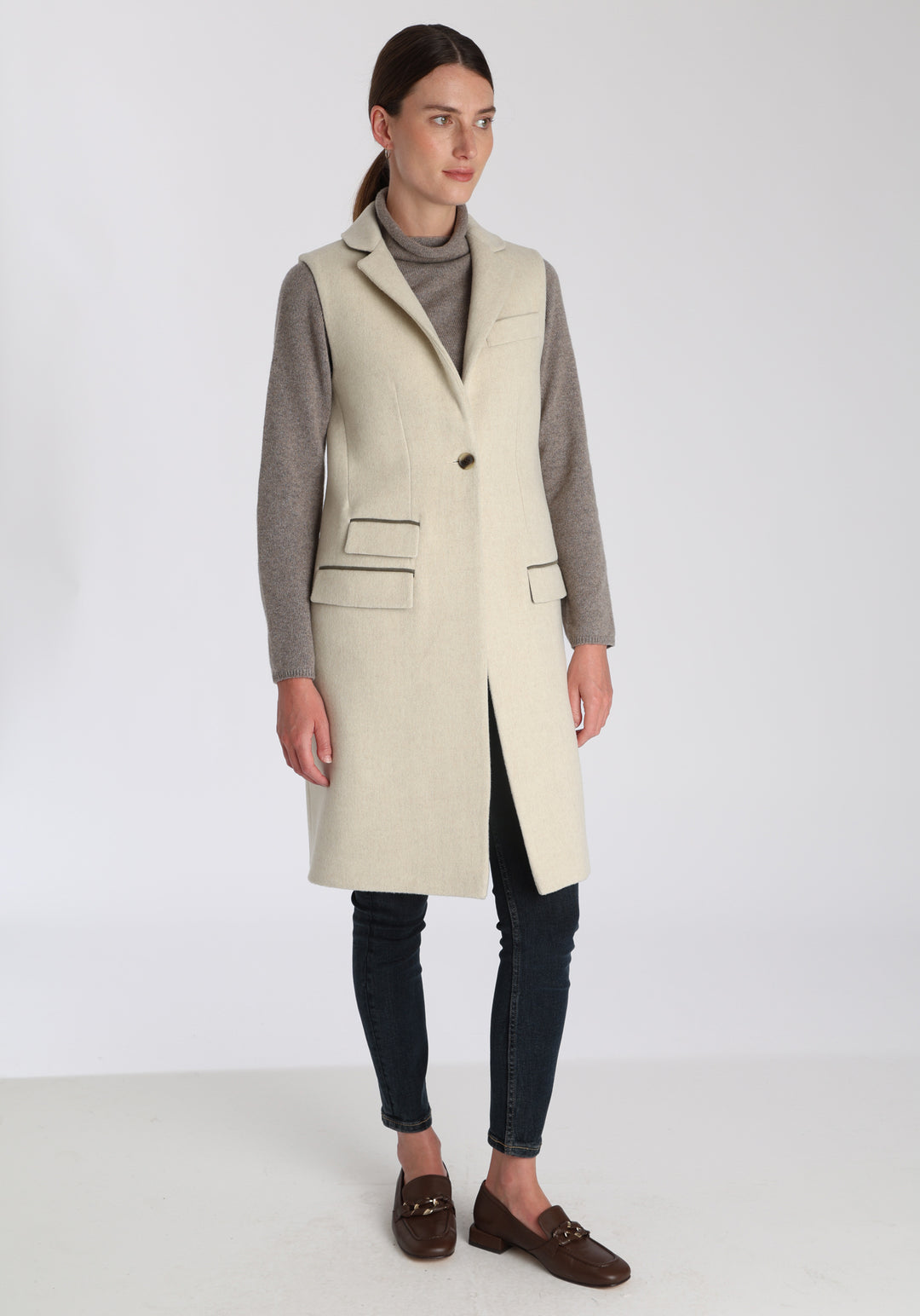 Model wearing the Kiekko waistcoat in cream merino wool with suede trim, a longline style featuring notched lapels, welt pockets, and a split back hem for ease of movement.