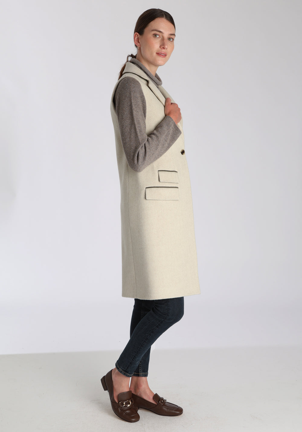 Model wearing the Kiekko waistcoat in cream merino wool with suede trim, a longline style featuring notched lapels, welt pockets, and a split back hem for ease of movement.