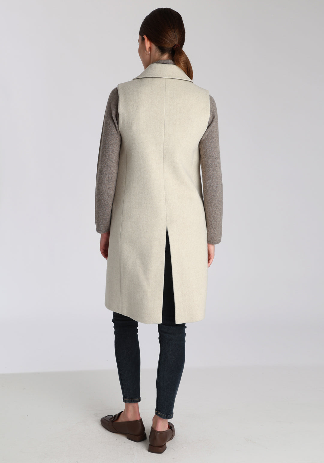 Model wearing the Kiekko waistcoat in cream merino wool with suede trim, a longline style featuring notched lapels, welt pockets, and a split back hem for ease of movement.