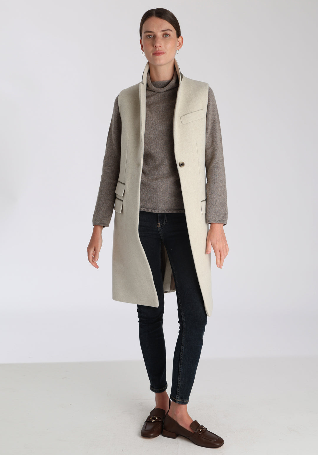 Model wearing the Kiekko waistcoat in cream merino wool with suede trim, a longline style featuring notched lapels, welt pockets, and a split back hem for ease of movement.