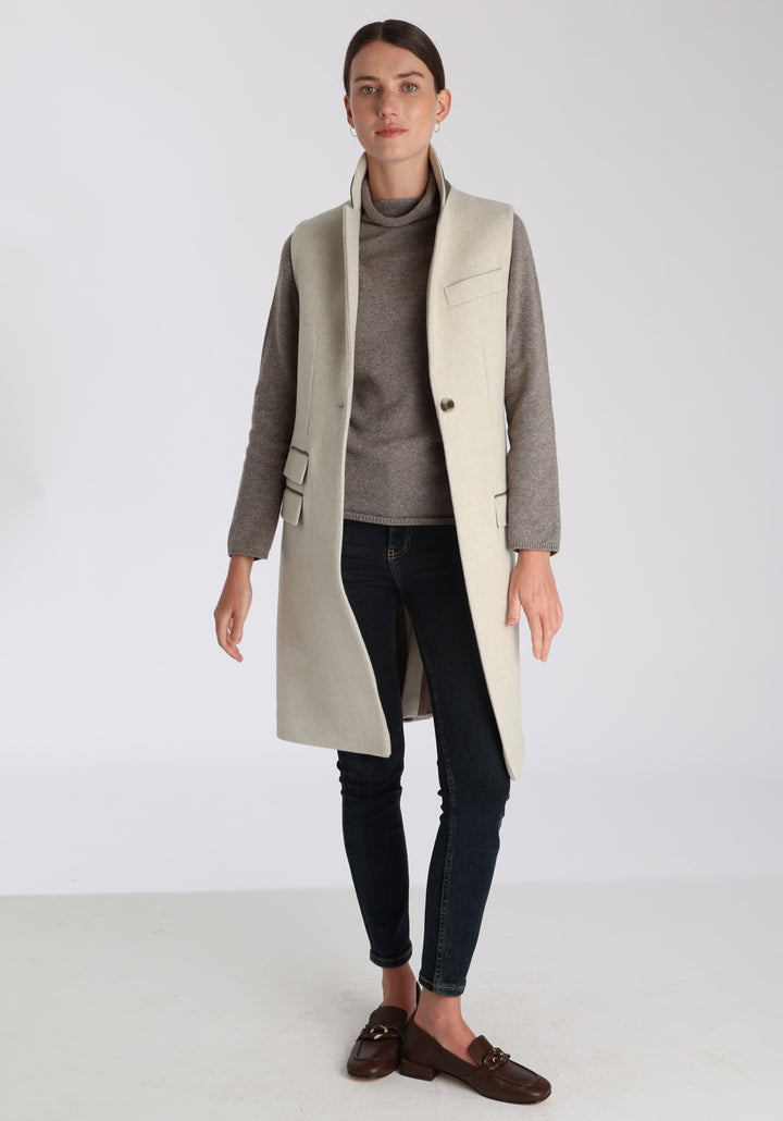 Model wearing the Kiekko waistcoat in cream merino wool with suede trim, a longline style featuring notched lapels, welt pockets, and a split back hem for ease of movement.