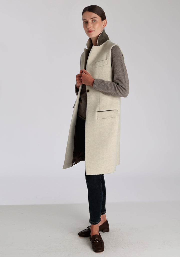 Model wearing the Kiekko waistcoat in cream merino wool with suede trim, a longline style featuring notched lapels, welt pockets, and a split back hem for ease of movement.