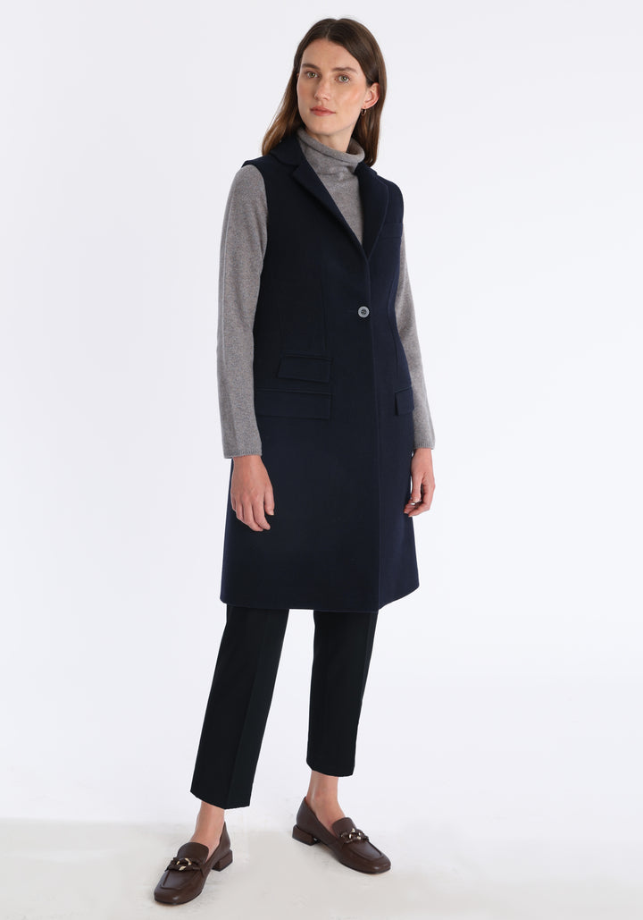 Model wearing the Kiekko waistcoat in navy merino wool with suede trim, a longline style featuring notched lapels, welt pockets, and a split back hem for ease of movement.