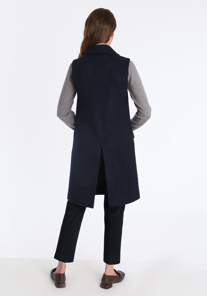 Model wearing the Kiekko waistcoat in navy merino wool with suede trim, a longline style featuring notched lapels, welt pockets, and a split back hem for ease of movement.