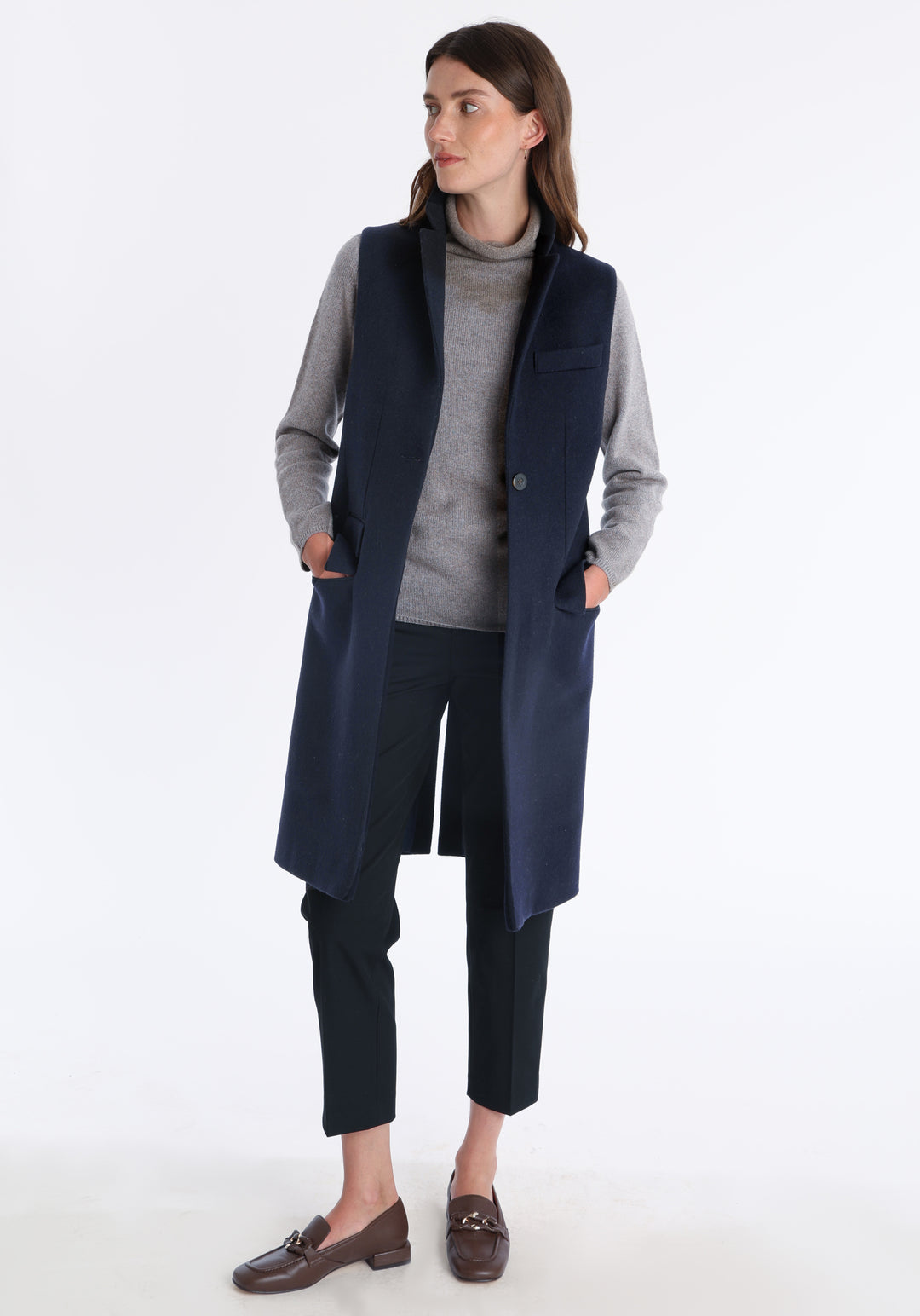 Model wearing the Kiekko waistcoat in navy merino wool with suede trim, a longline style featuring notched lapels, welt pockets, and a split back hem for ease of movement.
