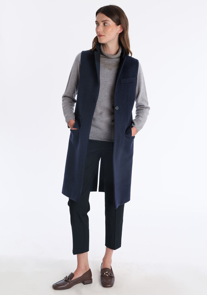 Model wearing the Kiekko waistcoat in navy merino wool with suede trim, a longline style featuring notched lapels, welt pockets, and a split back hem for ease of movement.