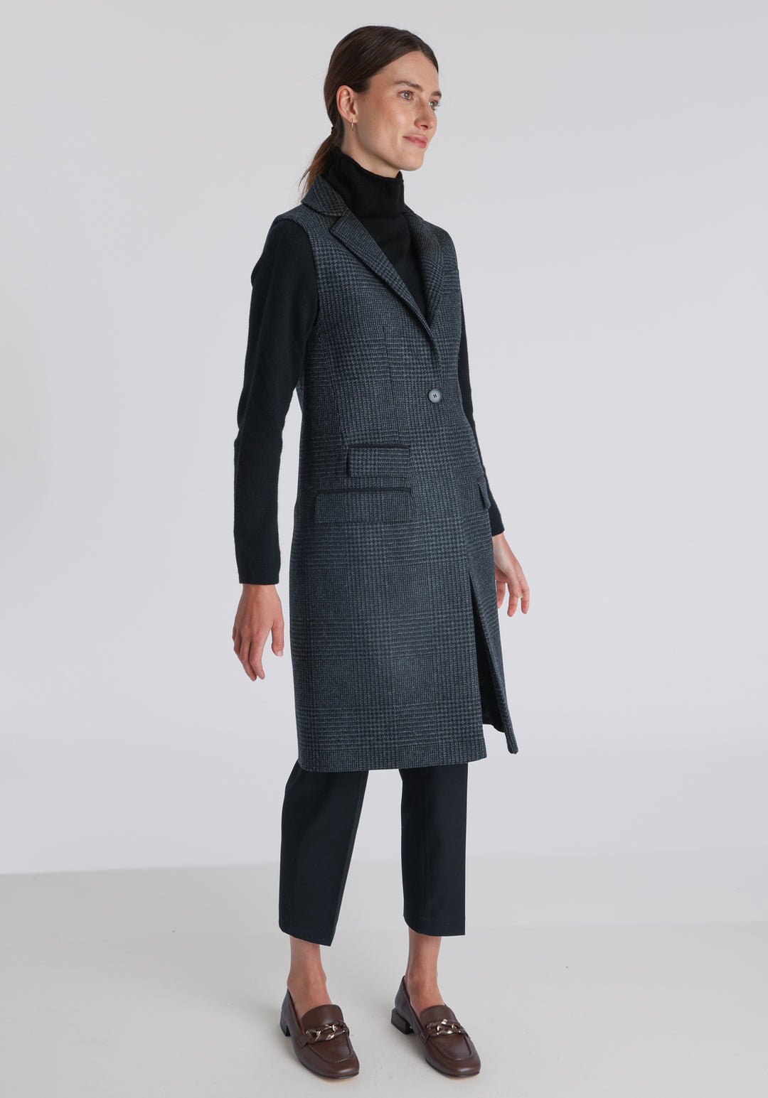 Model wearing the Kiekko waistcoat in black and grey Prince of Wales check with suede trim, a long line style featuring notched lapels, welt pockets, and a split back hem for ease of movement.