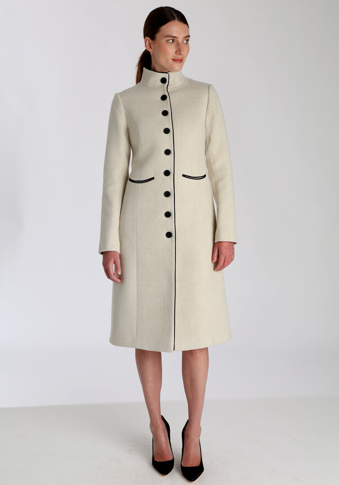 Model wearing the Opera coat in cream merino, an elegant, knee-length style with a high funnel neck, curved front pockets, and a slim, waist-contouring silhouette.