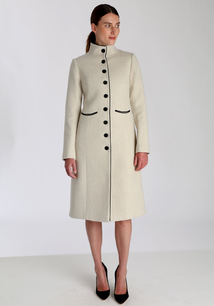 Model wearing the Opera coat in cream merino, an elegant, knee-length style with a high funnel neck, curved front pockets, and a slim, waist-contouring silhouette.