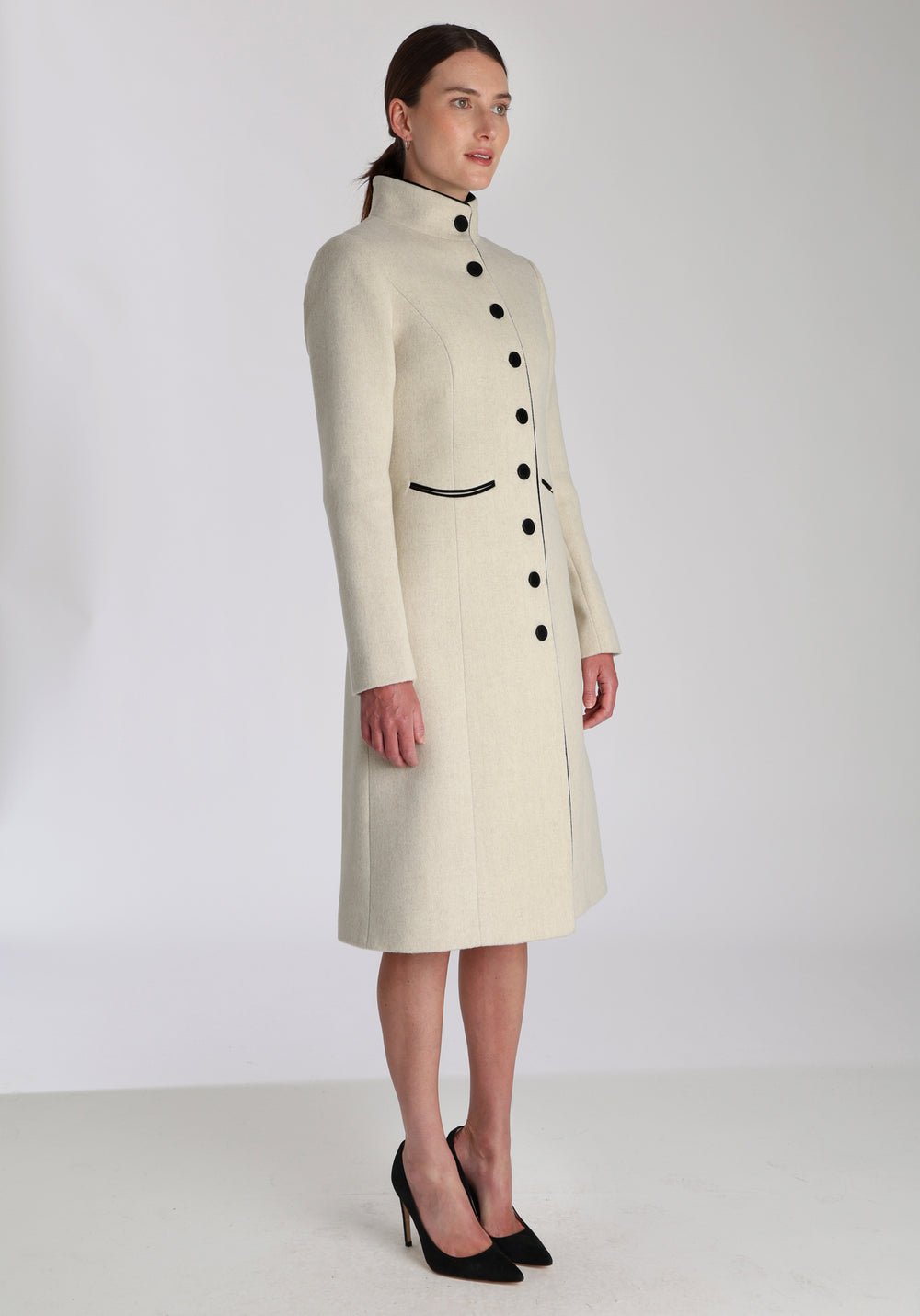 Model wearing the Opera coat in cream merino, an elegant, knee-length style with a high funnel neck, curved front pockets, and a slim, waist-contouring silhouette.