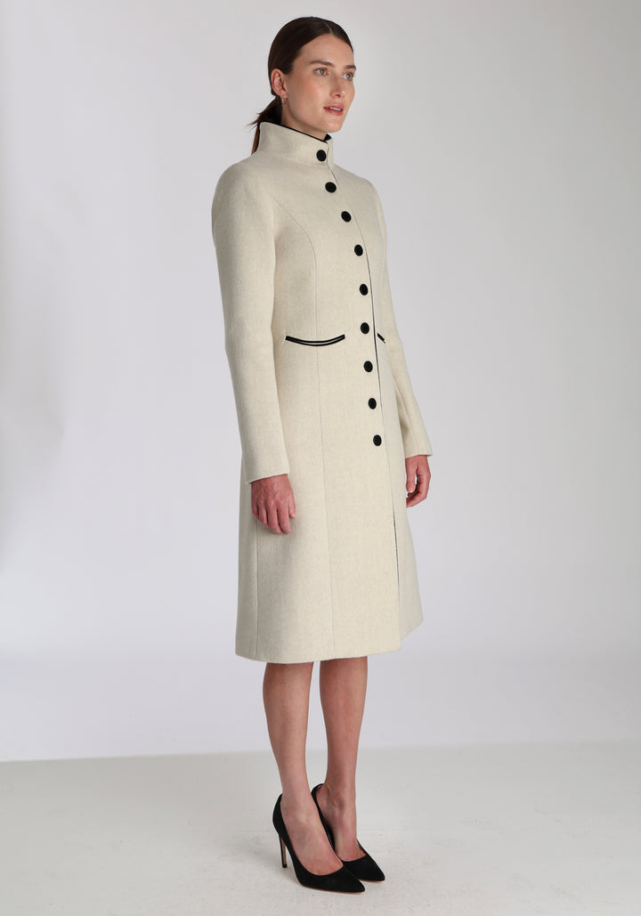 Model wearing the Opera coat in cream merino, an elegant, knee-length style with a high funnel neck, curved front pockets, and a slim, waist-contouring silhouette.