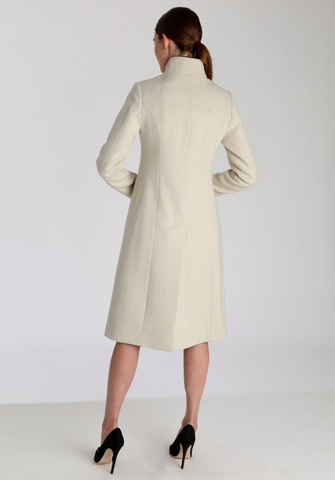 Model wearing the Opera coat in cream merino, an elegant, knee-length style with a high funnel neck, curved front pockets, and a slim, waist-contouring silhouette.