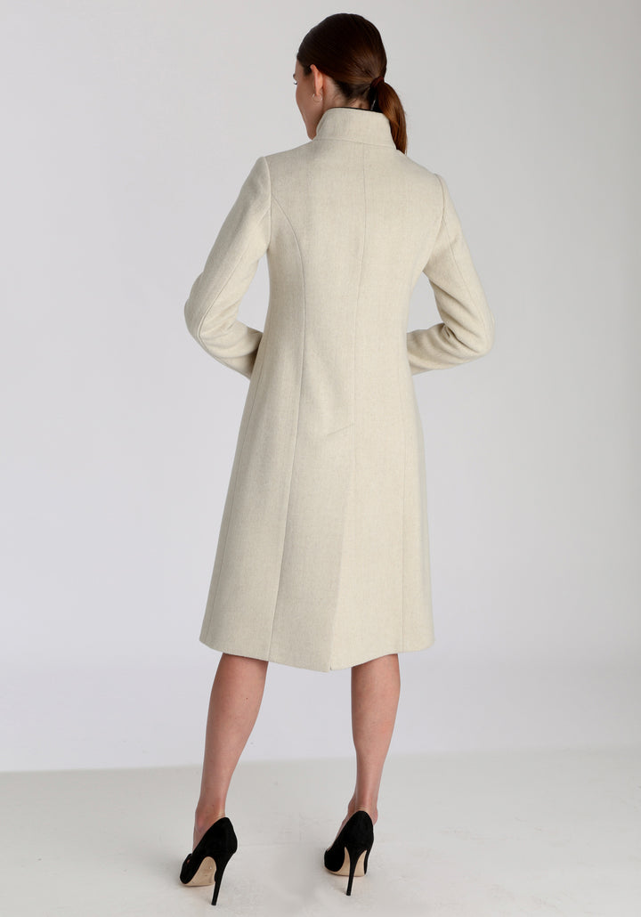 Model wearing the Opera coat in cream merino, an elegant, knee-length style with a high funnel neck, curved front pockets, and a slim, waist-contouring silhouette.