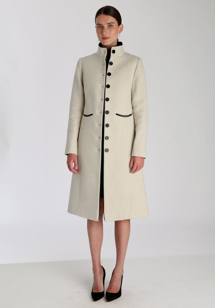 Model wearing the Opera coat in cream merino, an elegant, knee-length style with a high funnel neck, curved front pockets, and a slim, waist-contouring silhouette.