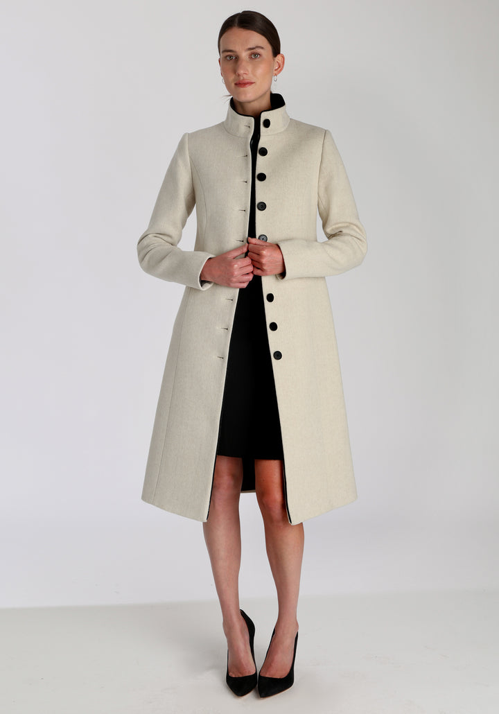 Model wearing the Opera coat in cream merino, an elegant, knee-length style with a high funnel neck, curved front pockets, and a slim, waist-contouring silhouette.