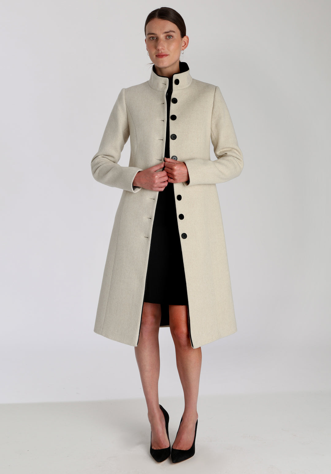 Model wearing the Opera coat in cream merino, an elegant, knee-length style with a high funnel neck, curved front pockets, and a slim, waist-contouring silhouette.