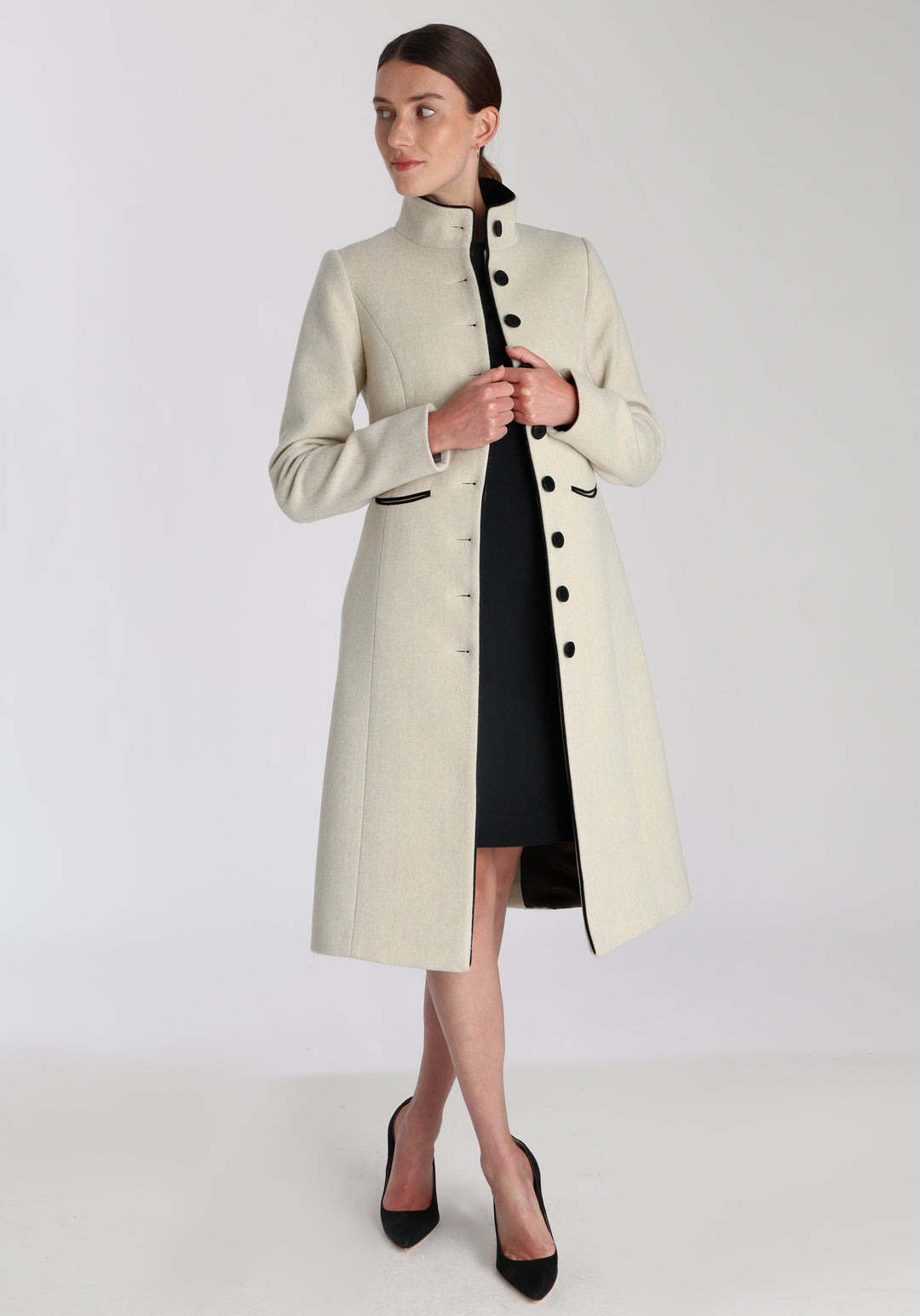 Model wearing the Opera coat in cream merino, an elegant, knee-length style with a high funnel neck, curved front pockets, and a slim, waist-contouring silhouette.