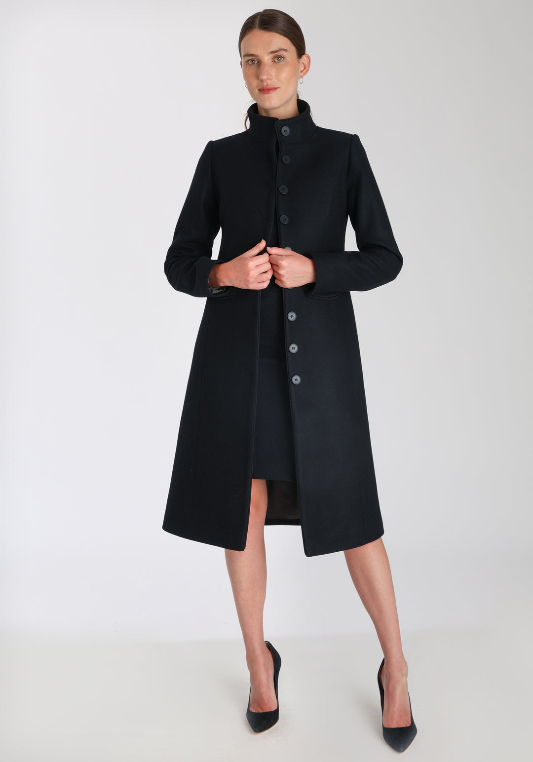 Model wearing the Opera coat in black cashmere blend, an elegant, knee-length style with a high funnel neck, curved front pockets, and a slim, waist-contouring silhouette.