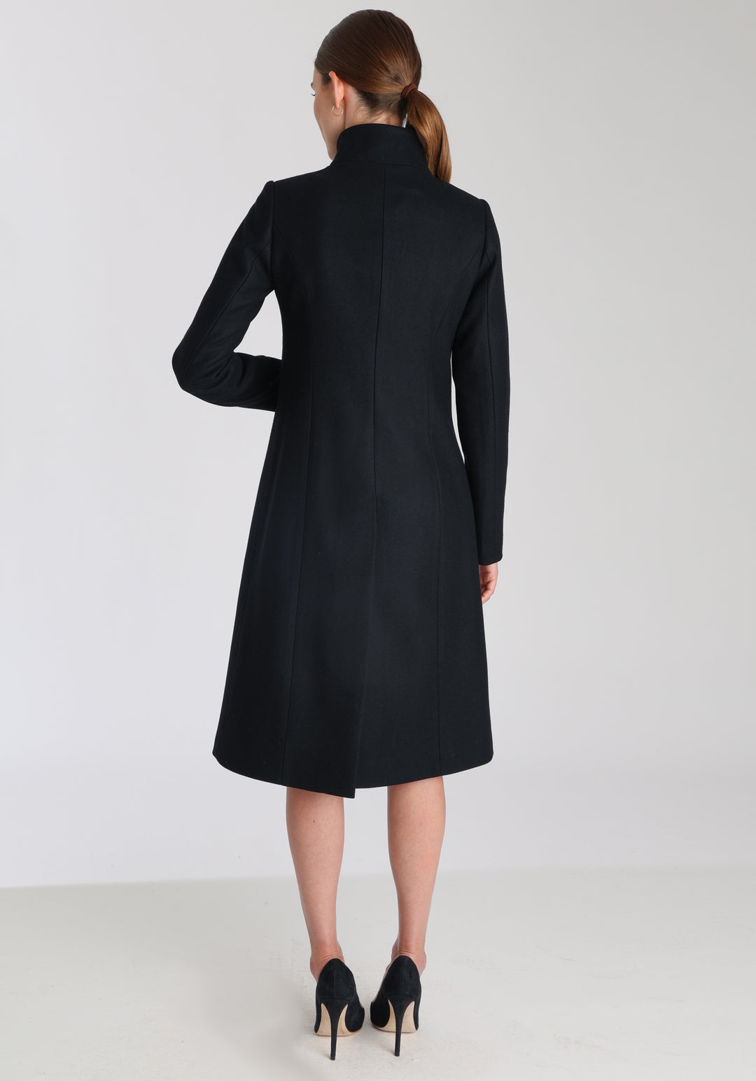 Model wearing the Opera coat in black cashmere blend, an elegant, knee-length style with a high funnel neck, curved front pockets, and a slim, waist-contouring silhouette.