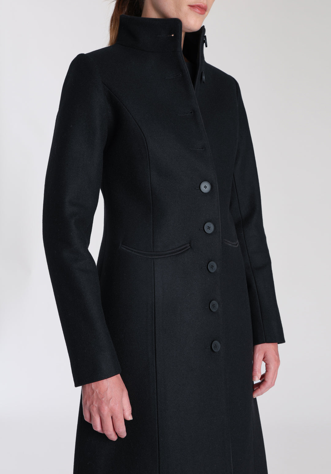 Model wearing the Opera coat in black cashmere blend, an elegant, knee-length style with a high funnel neck, curved front pockets, and a slim, waist-contouring silhouette.