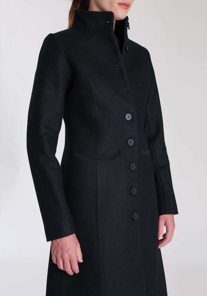 Model wearing the Opera coat in black cashmere blend, an elegant, knee-length style with a high funnel neck, curved front pockets, and a slim, waist-contouring silhouette.