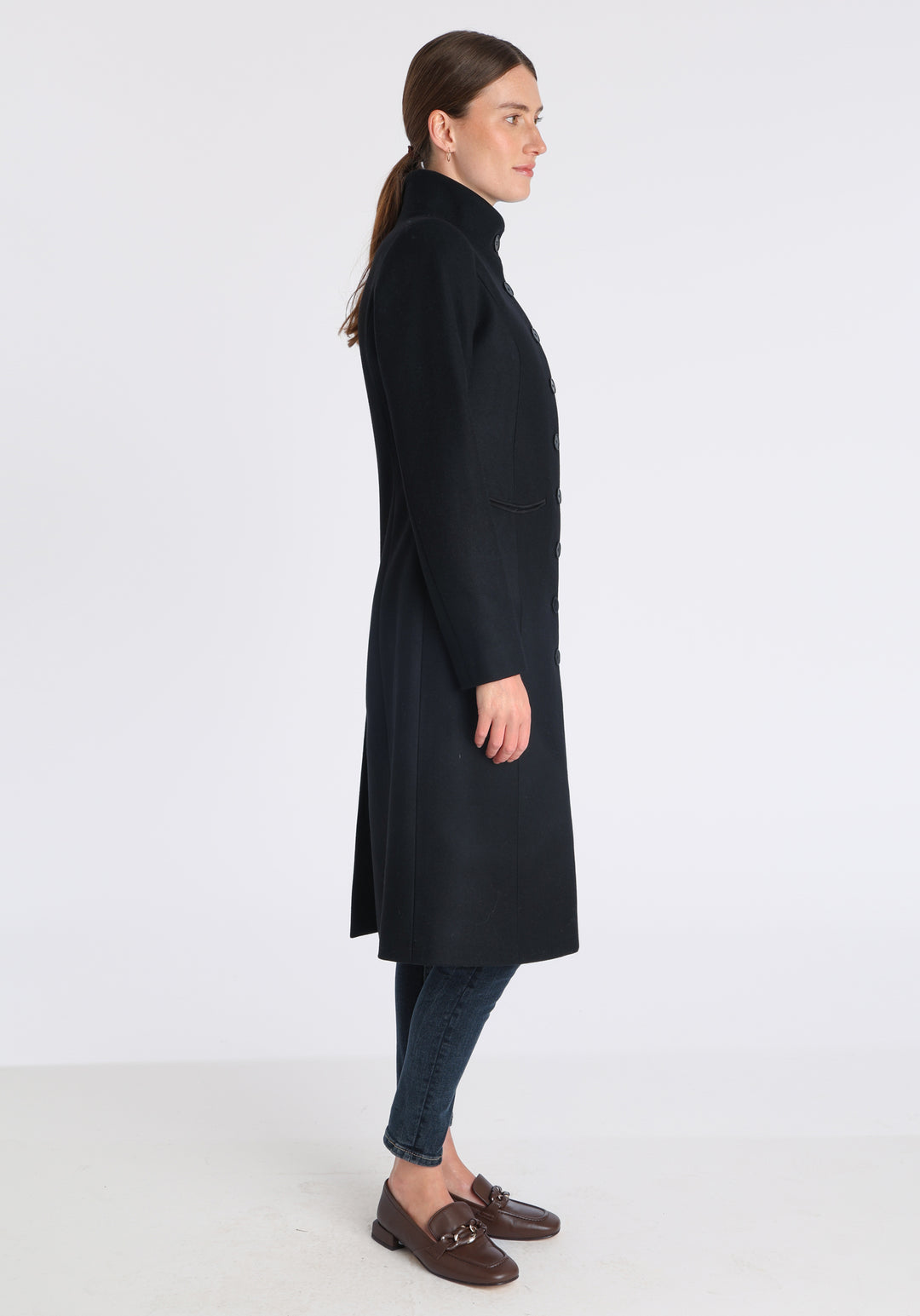 Model wearing the Opera coat in black cashmere blend, an elegant, knee-length style with a high funnel neck, curved front pockets, and a slim, waist-contouring silhouette.