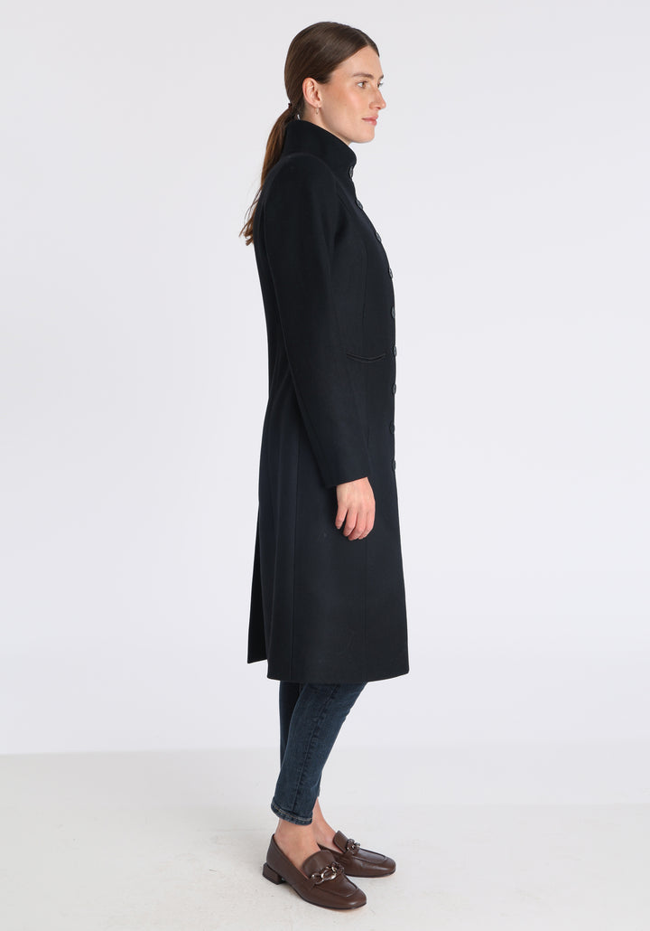 Model wearing the Opera coat in black cashmere blend, an elegant, knee-length style with a high funnel neck, curved front pockets, and a slim, waist-contouring silhouette.