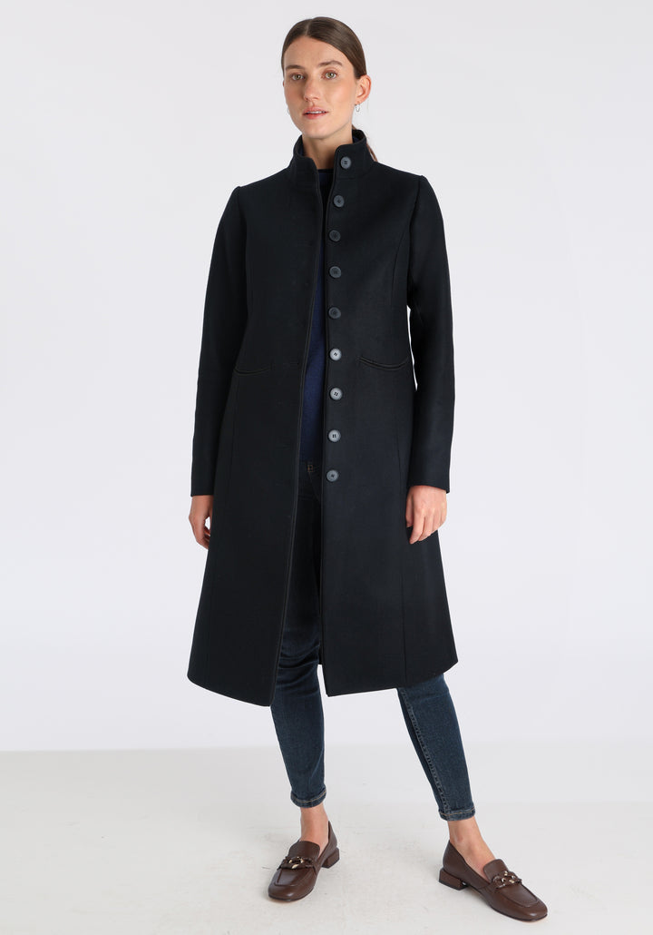 Model wearing the Opera coat in black cashmere blend, an elegant, knee-length style with a high funnel neck, curved front pockets, and a slim, waist-contouring silhouette.