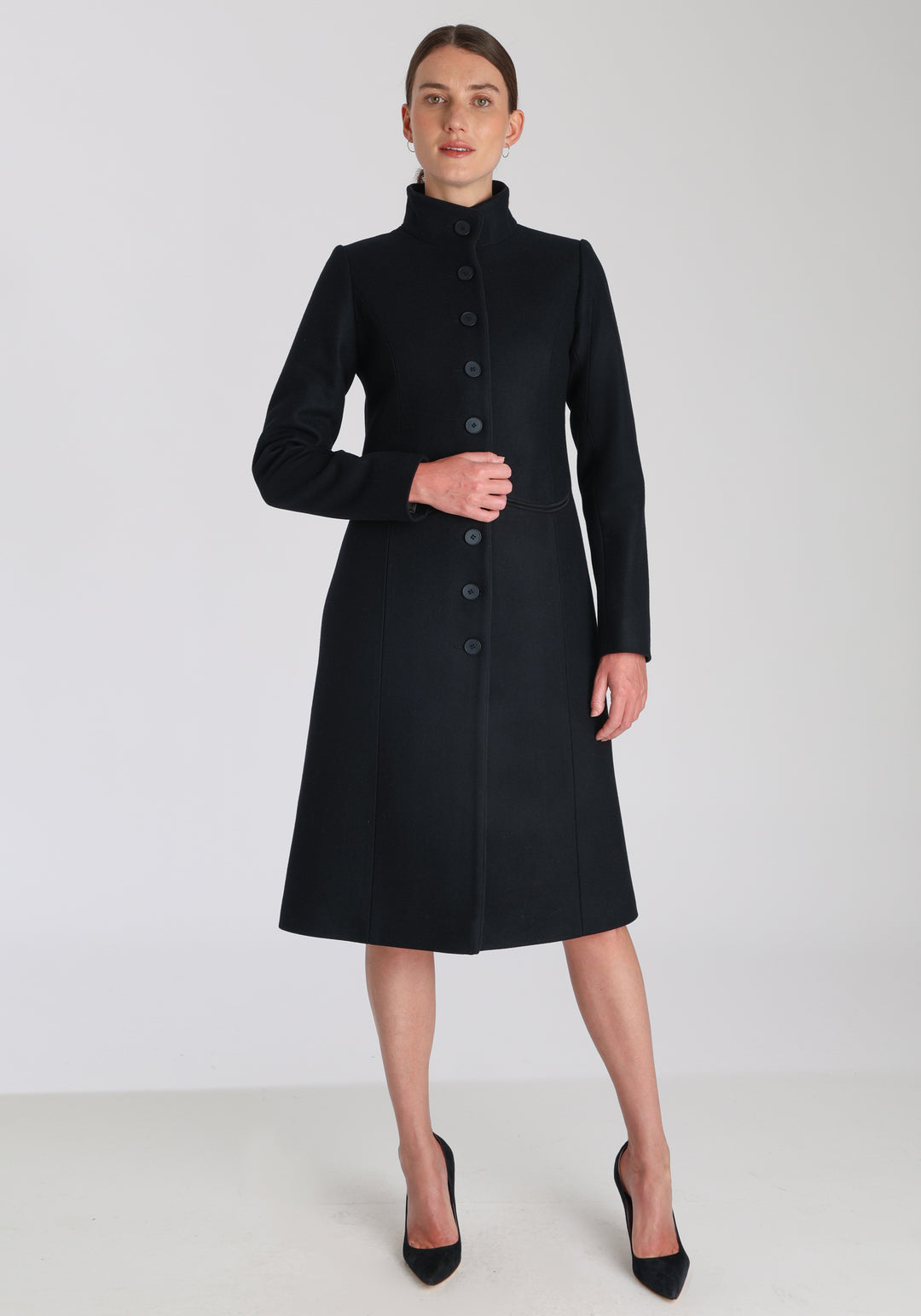 Model wearing the Opera coat in black cashmere blend, an elegant, knee-length style with a high funnel neck, curved front pockets, and a slim, waist-contouring silhouette.
