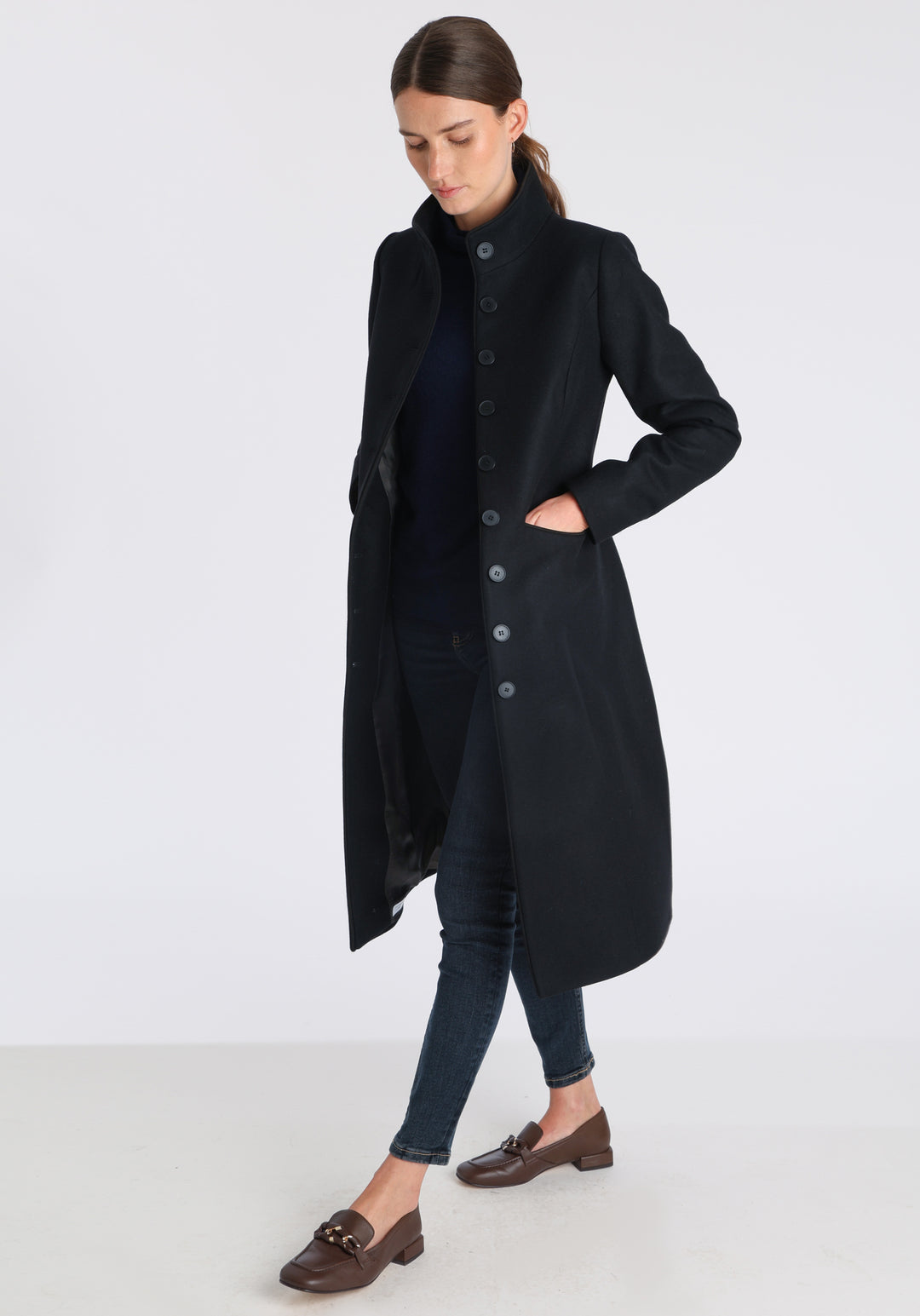 Model wearing the Opera coat in black cashmere blend, an elegant, knee-length style with a high funnel neck, curved front pockets, and a slim, waist-contouring silhouette.